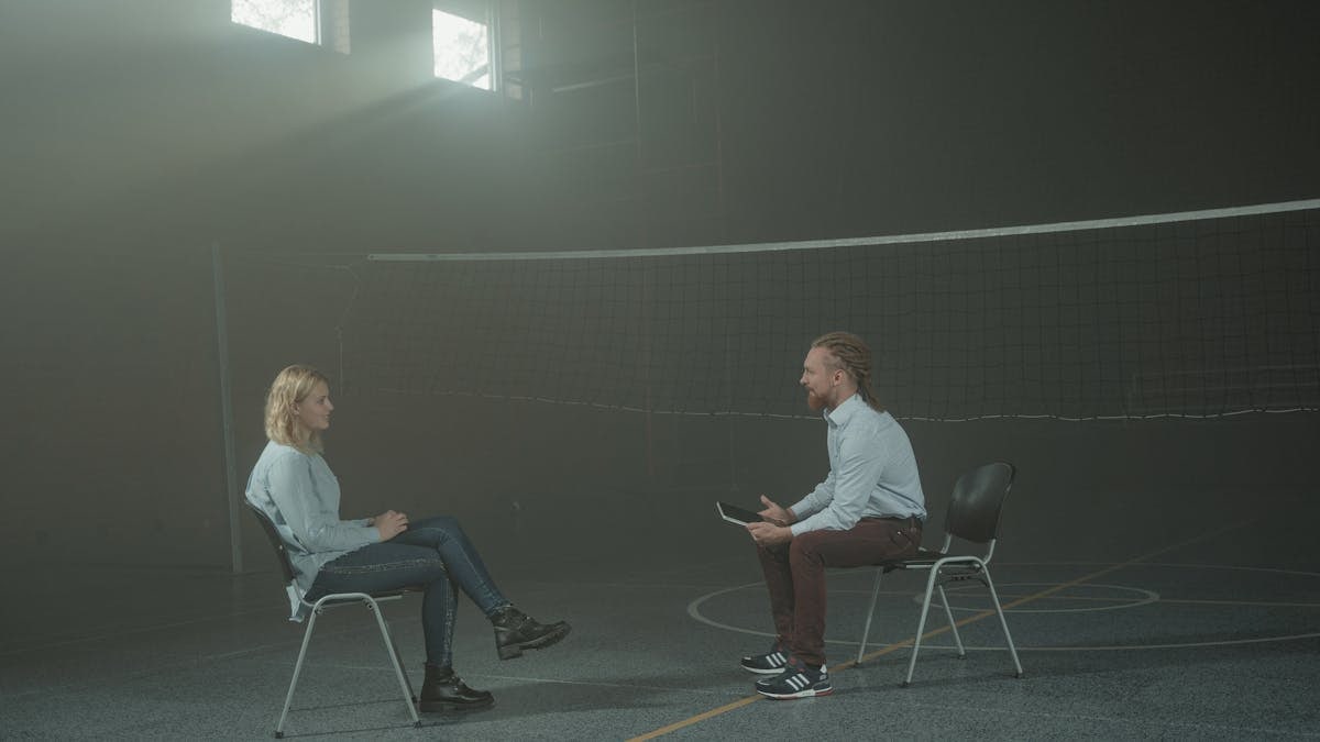 A man and a woman engage in a therapy session in a smoky indoor setting, sitting apart in a large room.